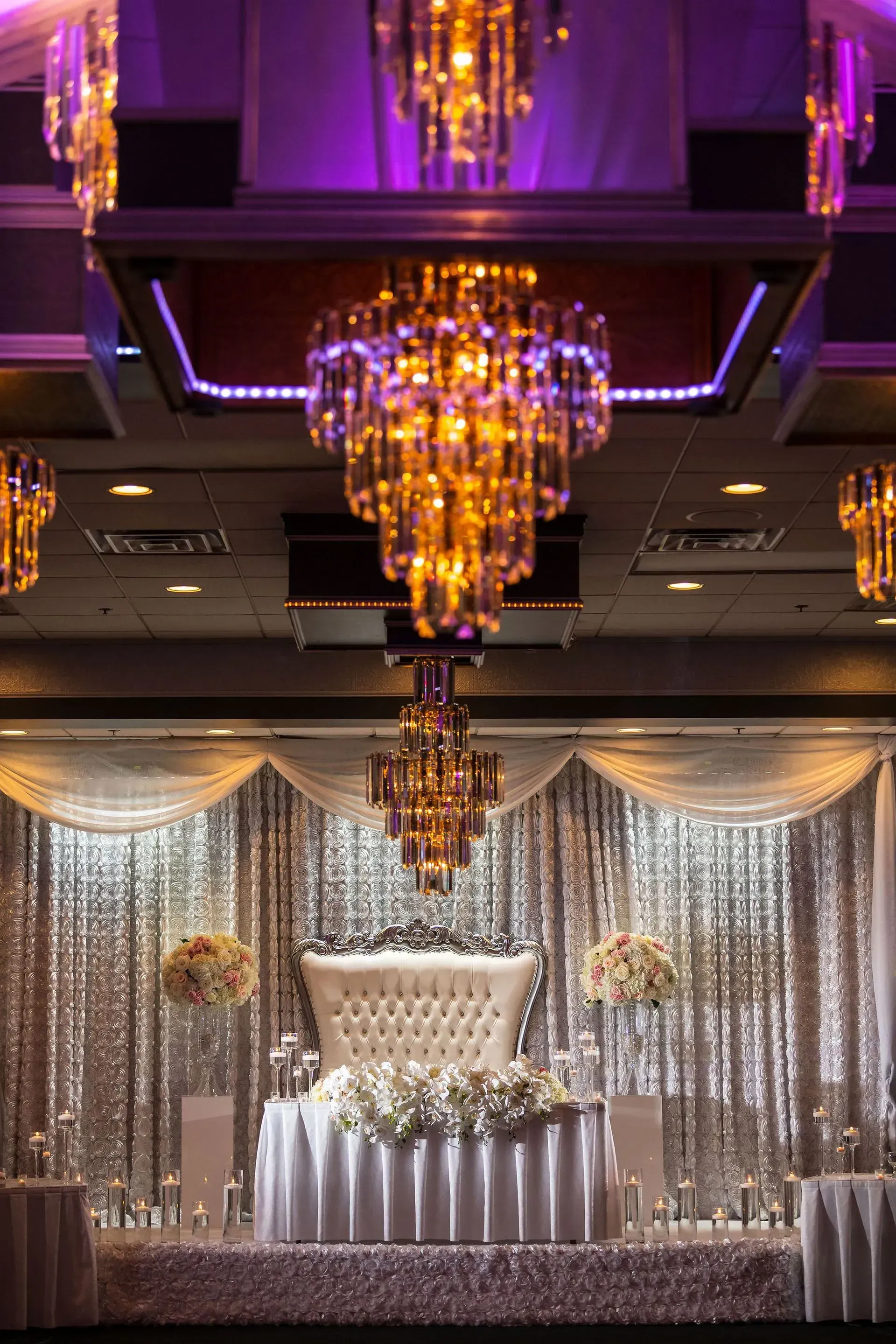 Purple-lit ballroom stage with crystal chandeliers, silver drapes, and a white floral display on a table