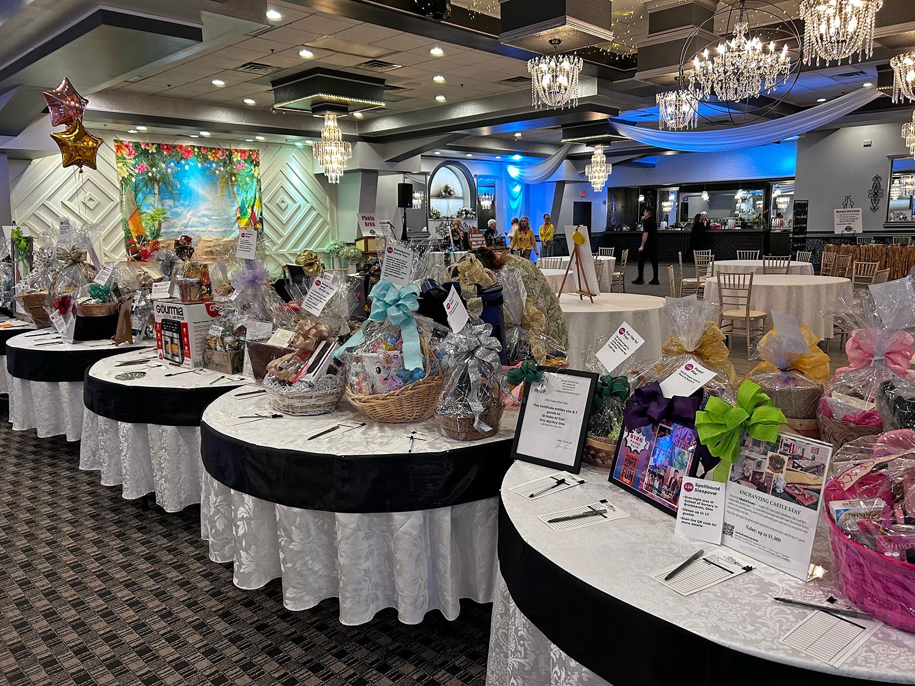 Decorated banquet hall with gift baskets displayed on round tables and chandeliers overhead.