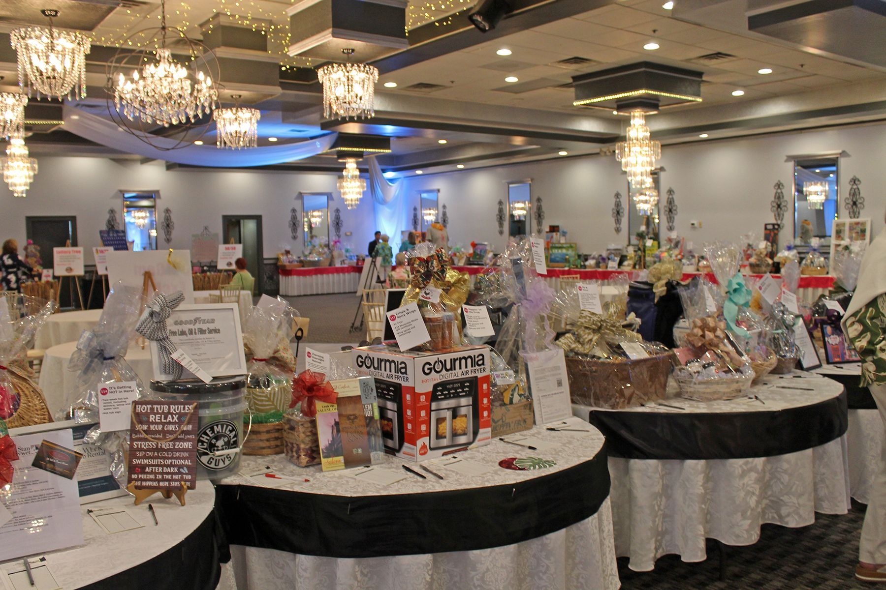 Decorated banquet hall with auction tables covered in gift baskets, bottles, and event prizes under chandeliers