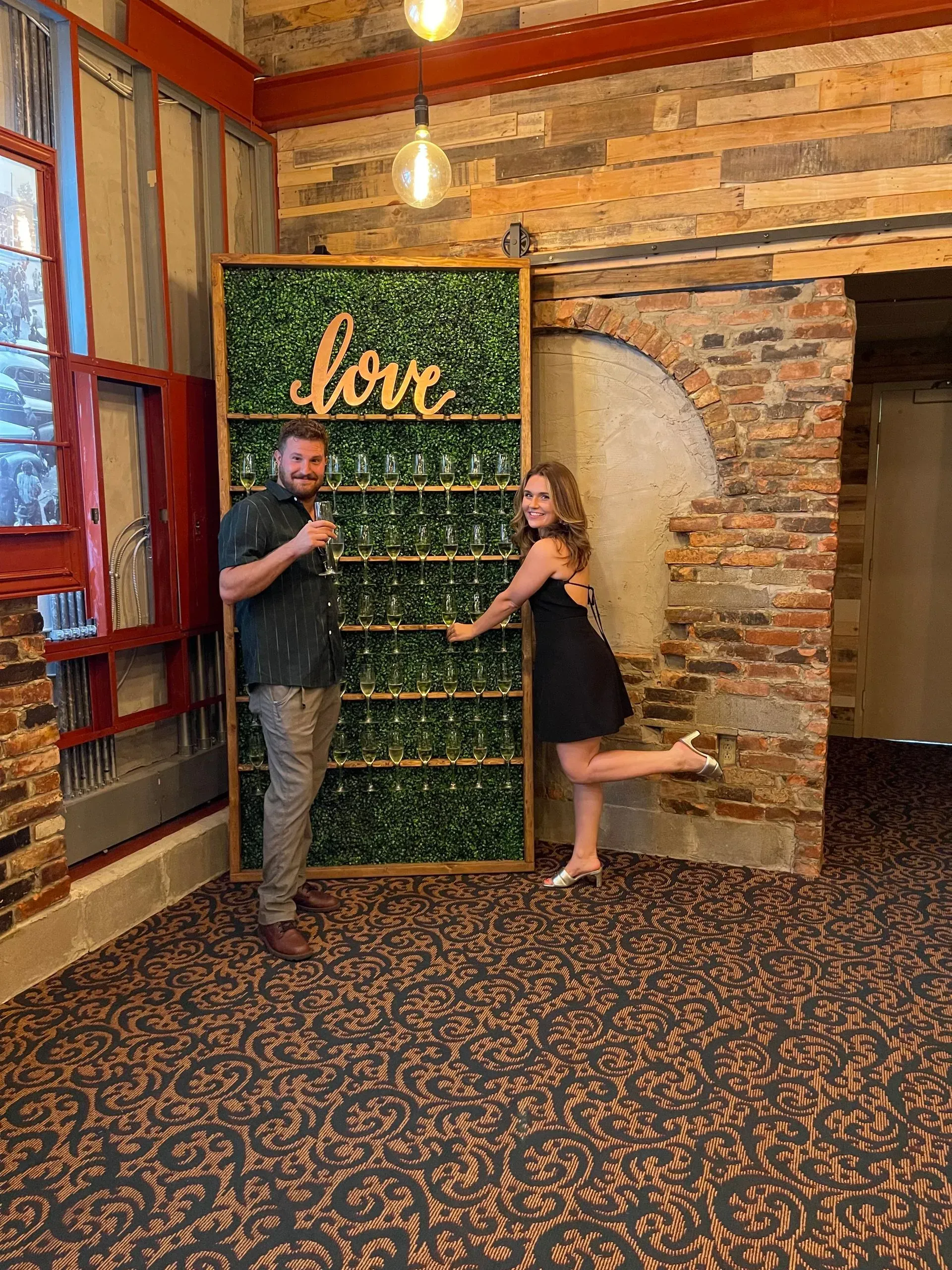 Two people posing beside a green floral wall with the word “love” in a rustic indoor setting