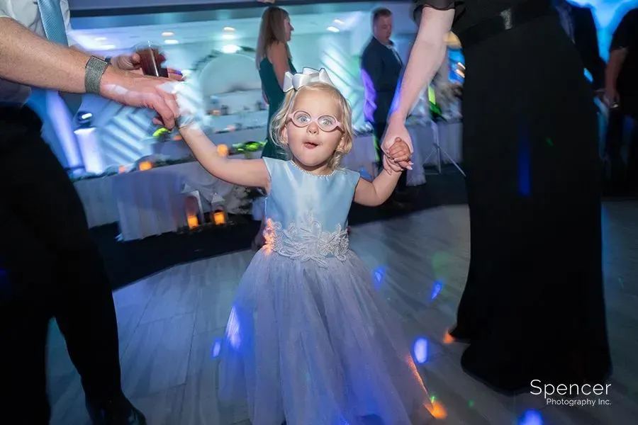 Young child in a white dress and glasses dancing at a party with blue lights, holding adults’ hands