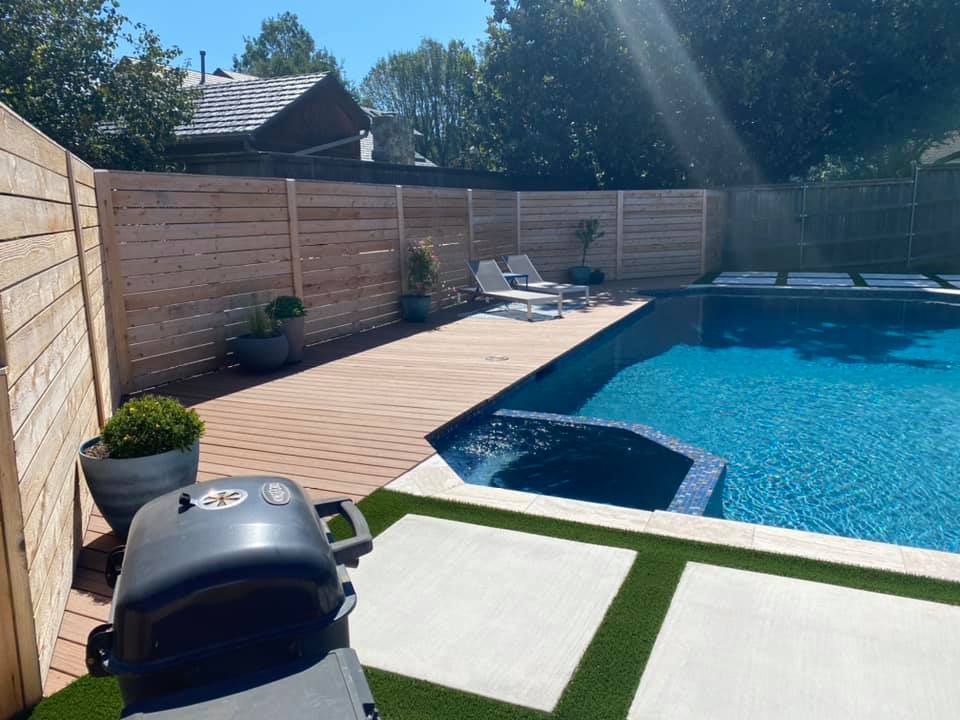 A large swimming pool is surrounded by a wooden fence and a grill.