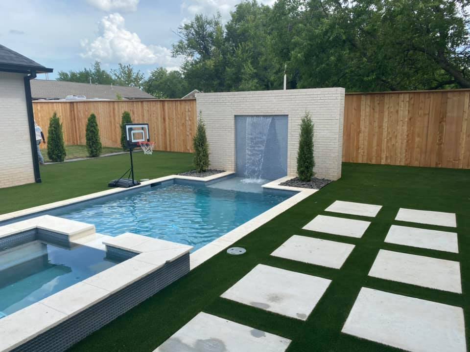 A swimming pool with a waterfall and a basketball hoop in the backyard of a house.