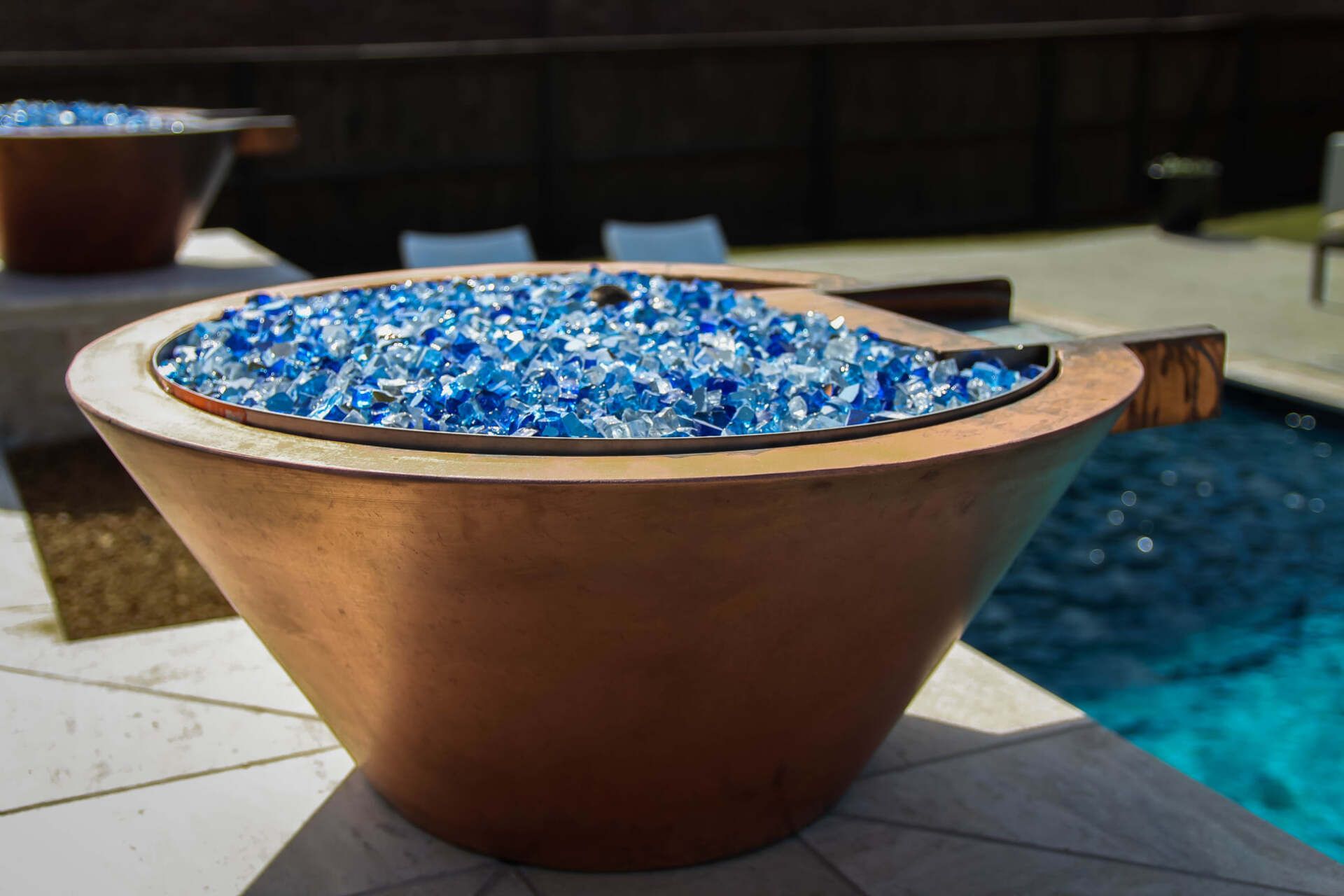 A fire pit filled with blue glass is sitting next to a pool.