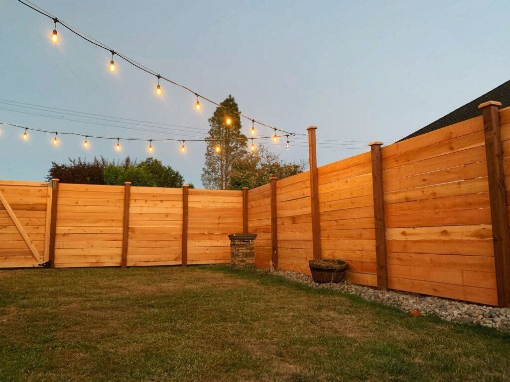 Wooden fence in a backyard with string lights overhead, grass lawn.