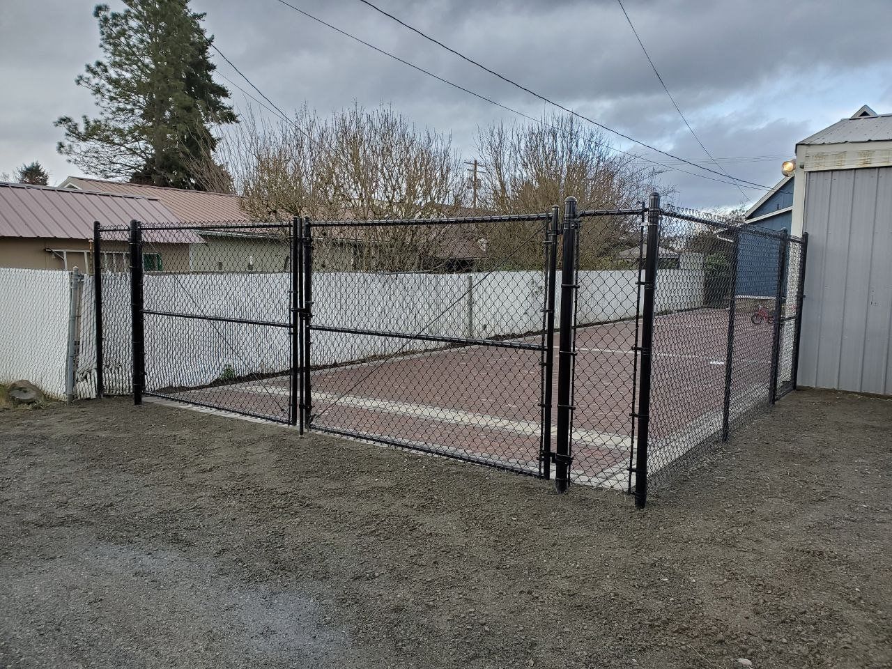 Black chain-link fence encloses a dirt area, gates open, gravel ground.