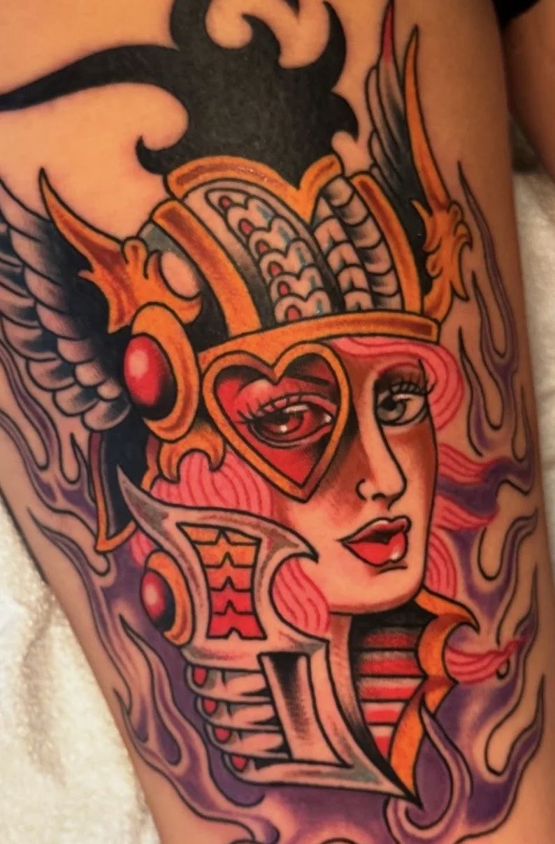 Colorful tattoo of a stylized face with red eyes, pink skin, and ornate headdress on skin background.