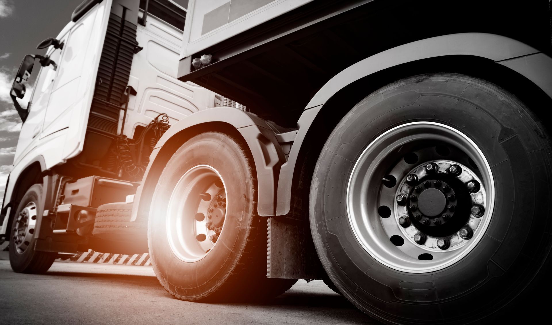 A close-up, low-angle view of the large wheels and side panel of a white semi-truck on a road, with a bright light flare.