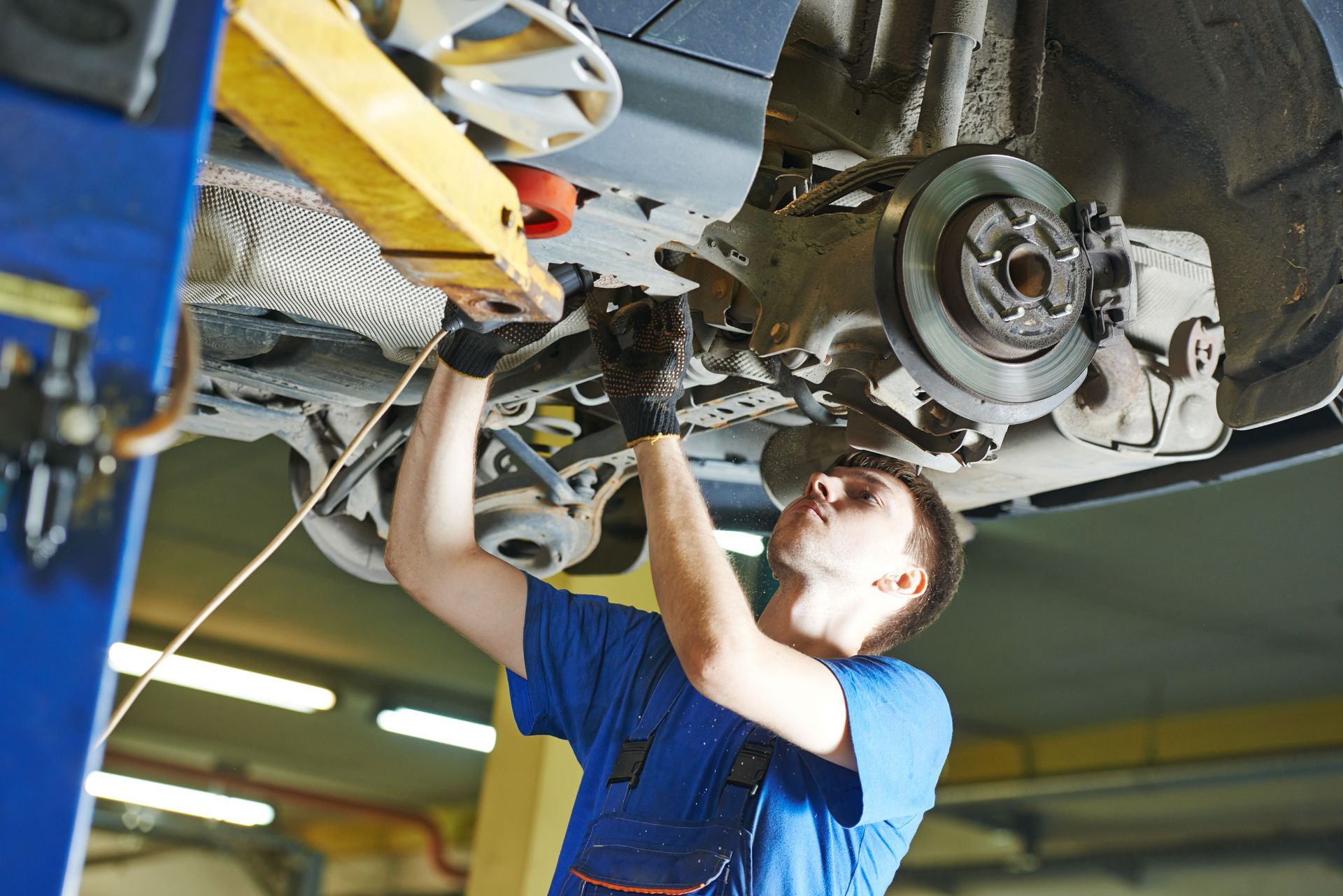 A mechanic in a blue uniform works on the underside of a car raised on a lift in a workshop.