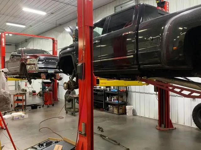 Two pickup trucks, one red and one black, are elevated on hydraulic lifts inside a bright automotive repair shop.