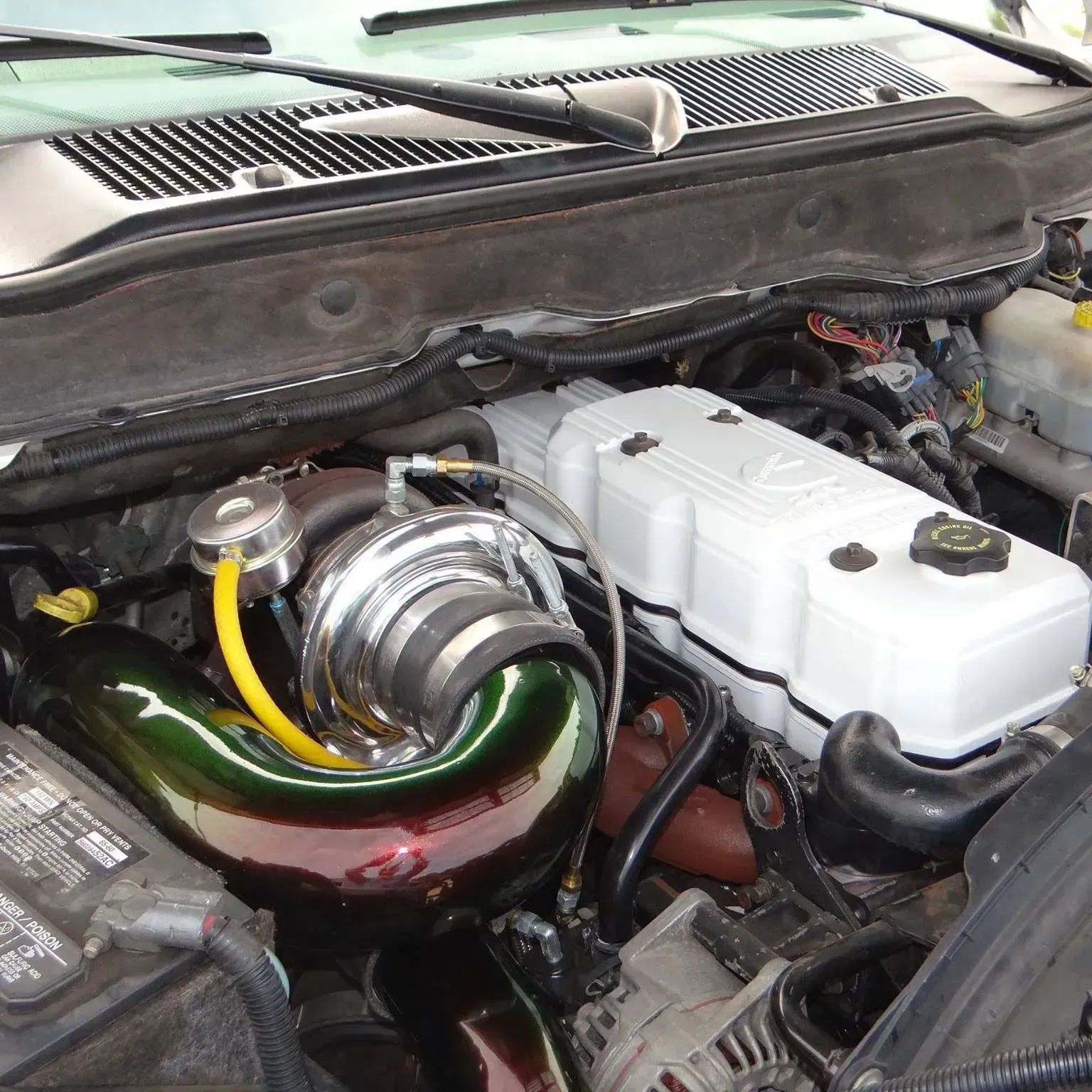 Engine bay of a truck featuring a prominent iridescent green turbocharger intake pipe and a white valve cover.