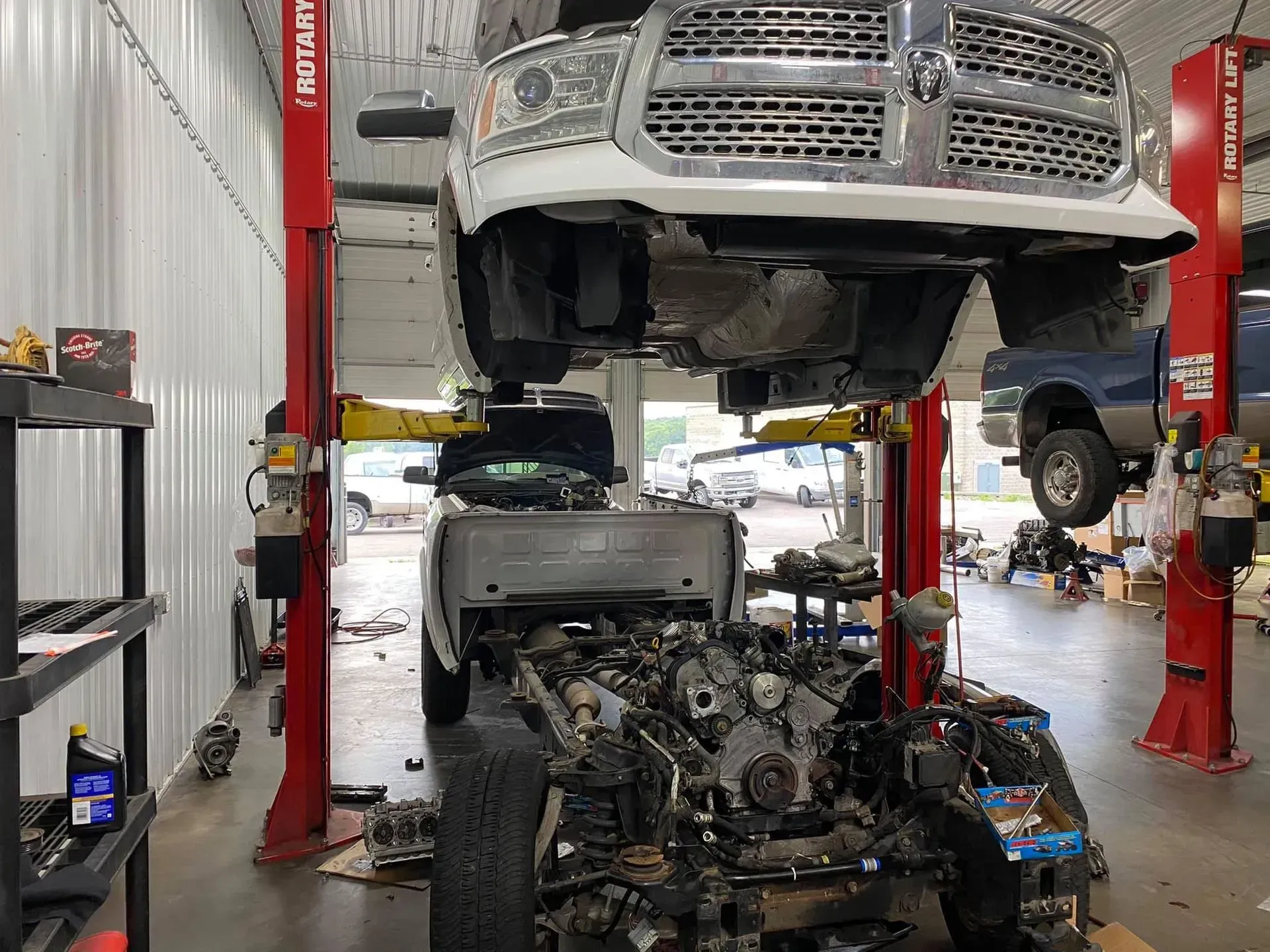 A white truck undergoing a chassis-cab separation on a red automotive lift inside a repair shop.