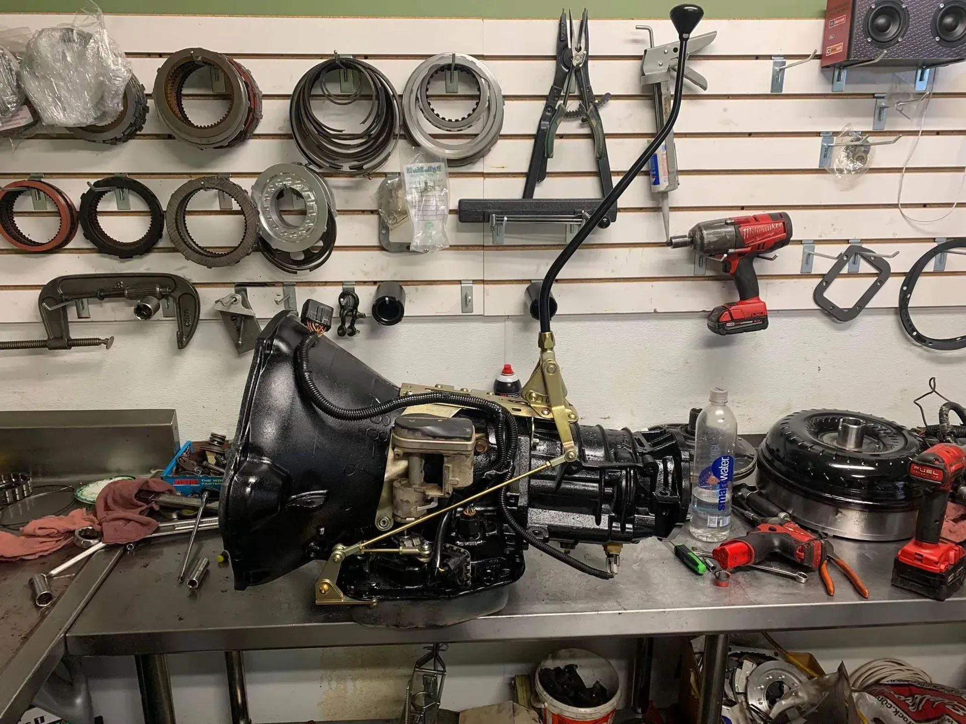 A disassembled car transmission with a tall gear shifter sitting on a metal workbench in a professional mechanic shop.