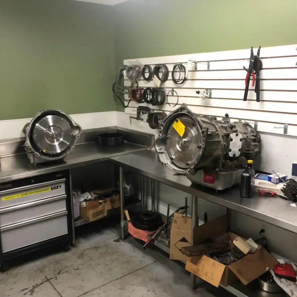 Two automotive transmissions sit on a stainless steel workbench in a shop, with tools hung on a wall panel behind them.