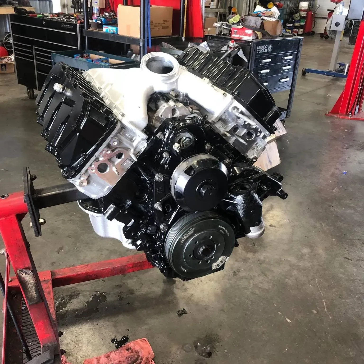 A freshly painted black and silver V8 engine sits mounted on a red metal engine stand inside a repair shop.