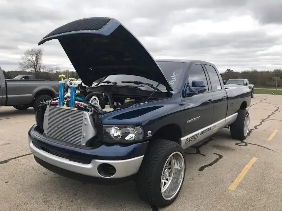A blue Dodge Ram pickup truck with its hood open, parked on asphalt with a large exposed radiator and custom wheels.