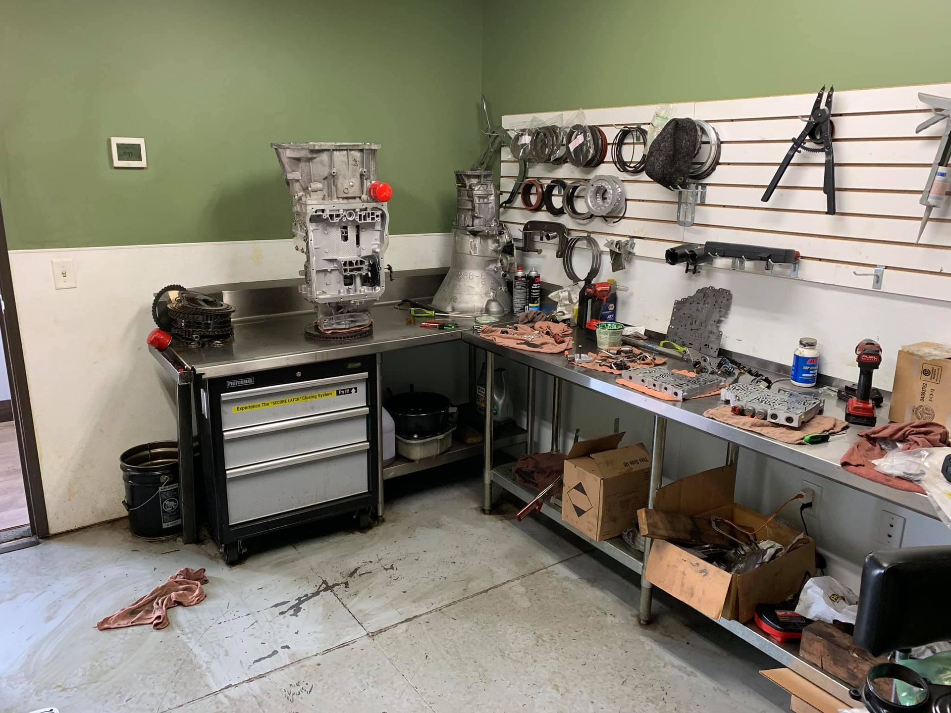 A workshop featuring an automotive transmission on a steel workbench and tools organized on a wall-mounted slat board.
