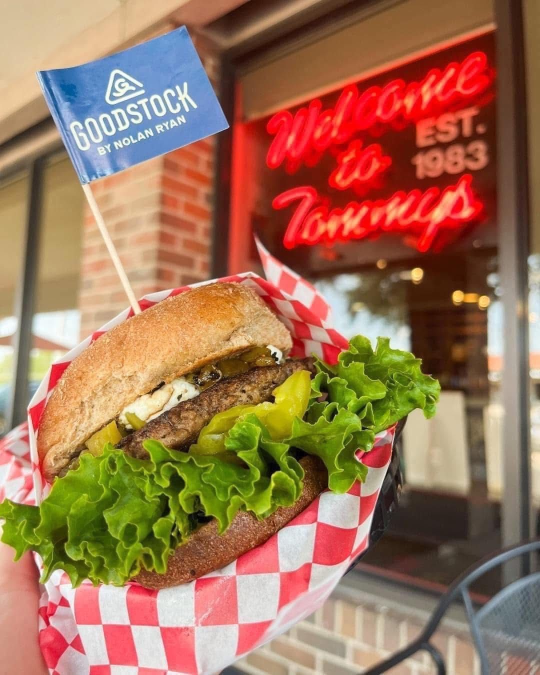 Hamburger with lettuce and pickles, held in checkered paper, with neon Tommy's sign in the background.