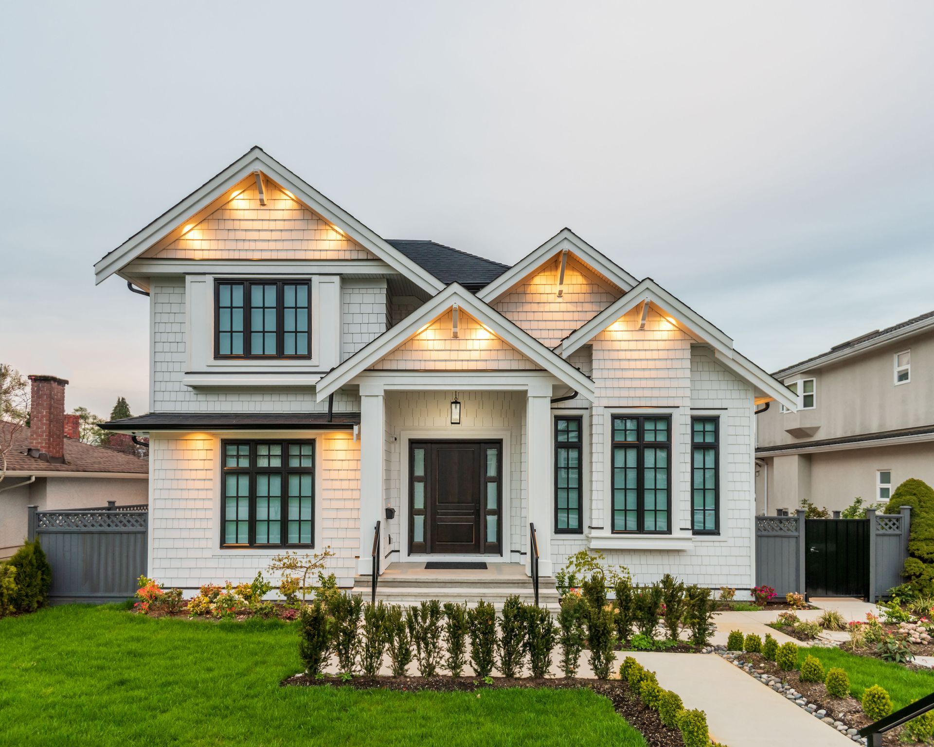 White two-story house with black windows, lit entry, green lawn, and pathway leading to the front door.