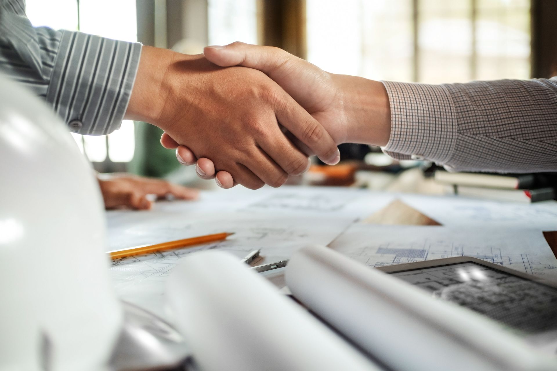 Two people shaking hands over blueprints in an office setting.