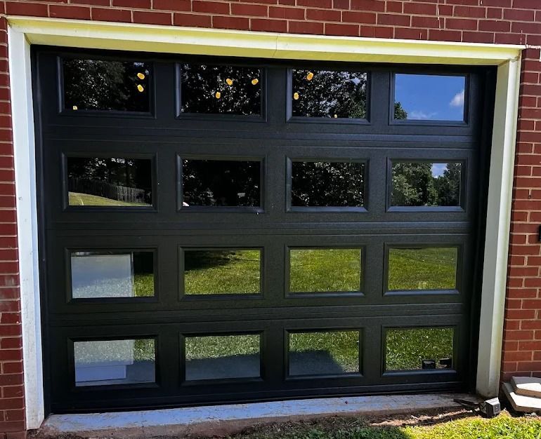 Black garage door with rectangular windows reflecting green grass and sky, framed by brick and white trim.