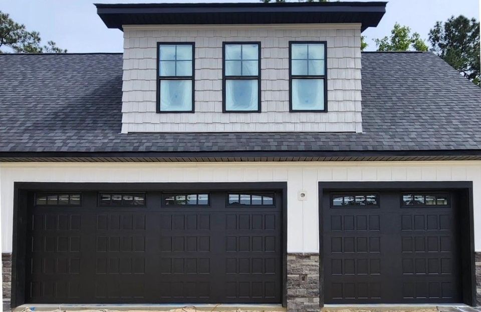Black garage doors with windows, stone accents, and a gray shingled facade. Three windows sit above.