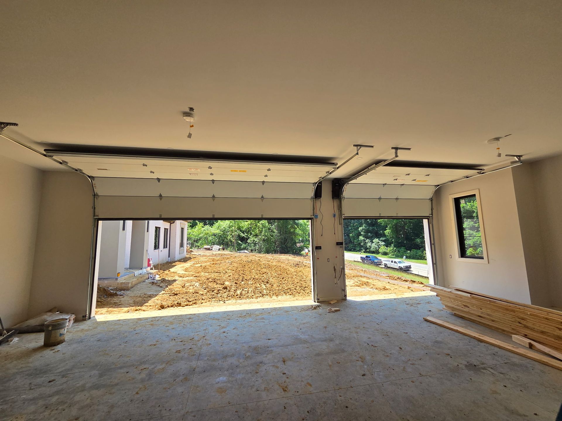 Interior of a garage under construction with open bays, a pile of wood, and greenery outside.