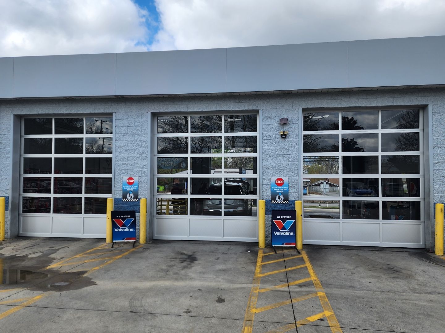 Three garage bays with glass doors at a service center. Blue building exterior, yellow lane markings, and two advertising displays are present.