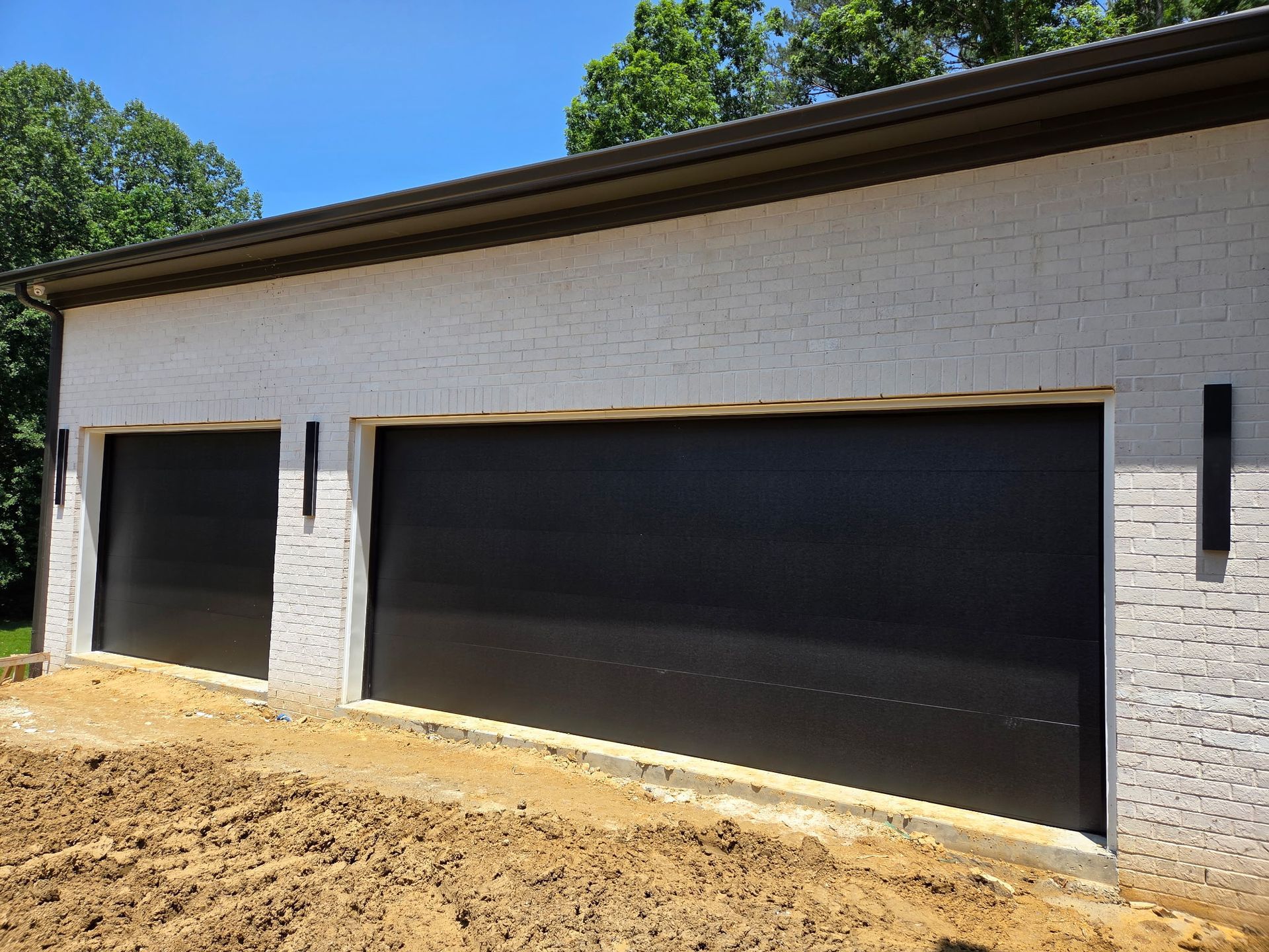Black garage doors in brick building. Brown earth in front. Green trees and blue sky in background.