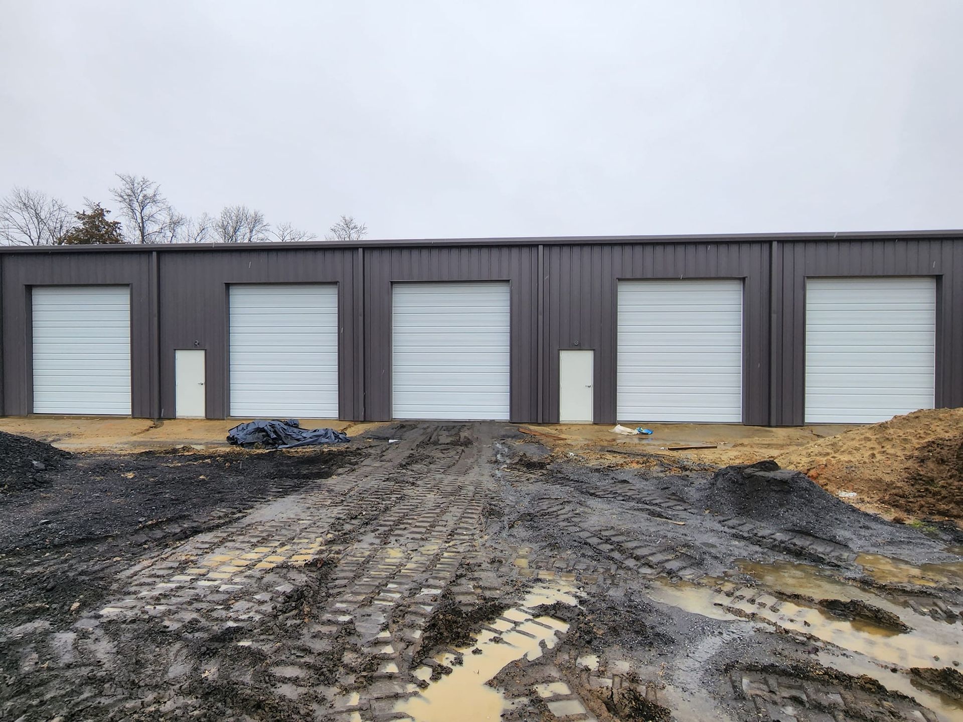 Row of brown storage units with white garage doors on a muddy construction site.