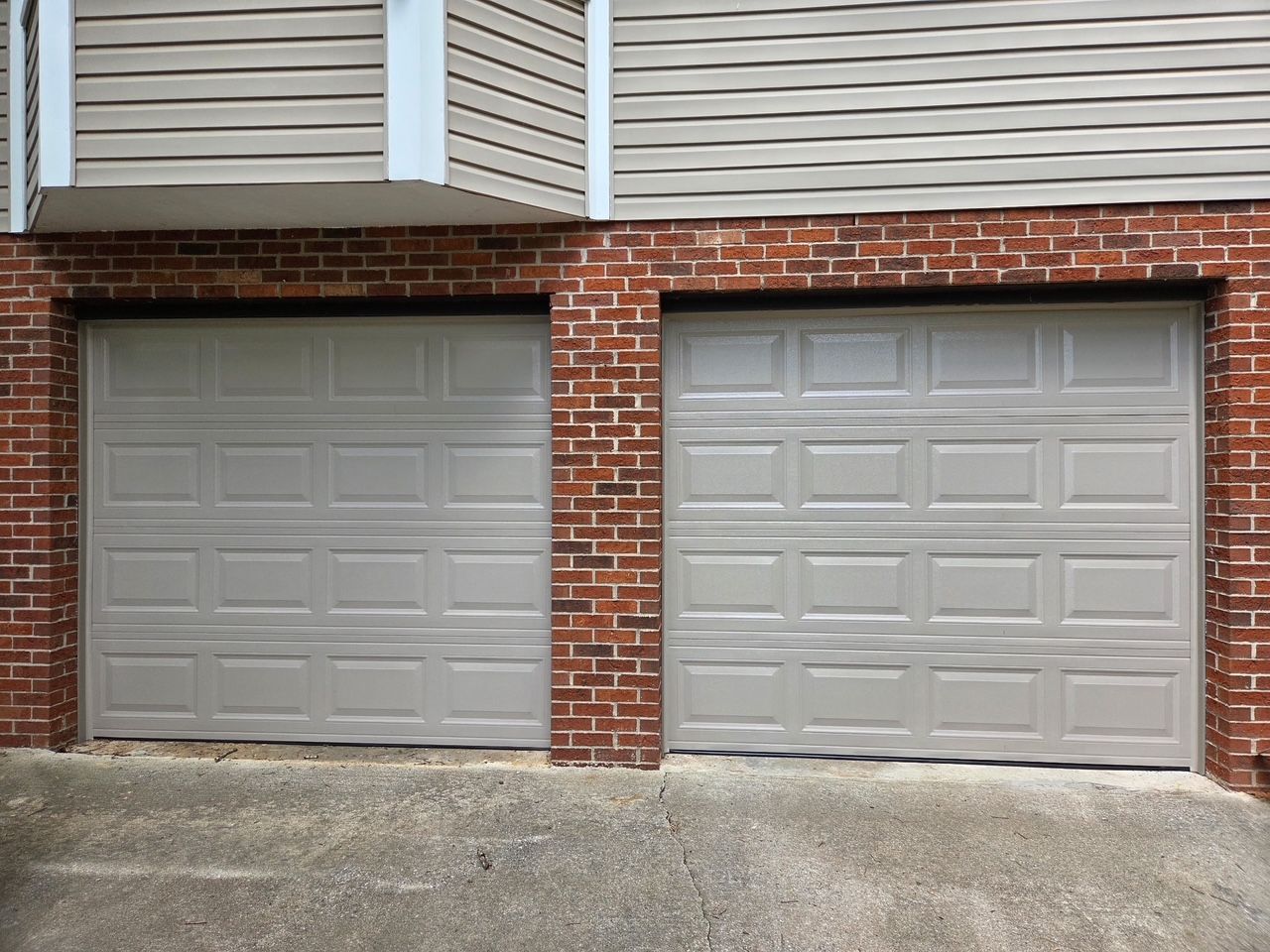 Two closed gray garage doors with red brick surrounding, beneath light brown siding.