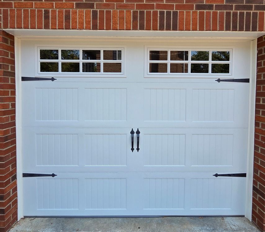 White garage door with windows and decorative black hardware against a brick wall.