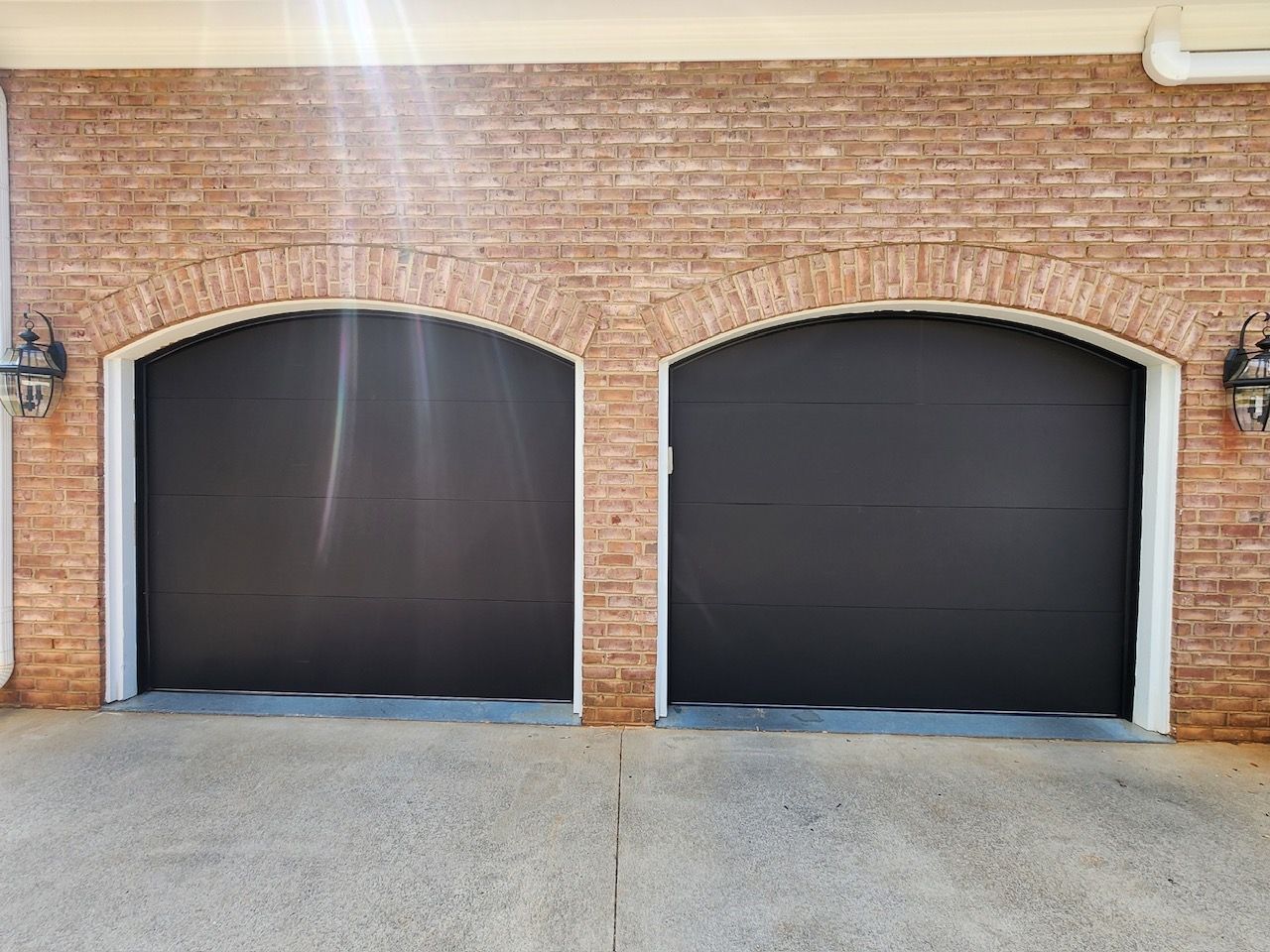 Two closed black garage doors under brick archways, set in a brick wall, with concrete driveway.