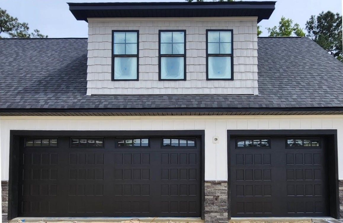 Two-car garage with dark brown doors, stone accents, and three windows in a gray-shingled gable.