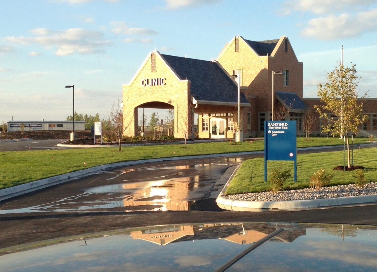 A tan brick medical clinic building with a covered entrance and blue signage on a sunny, grassy lot.