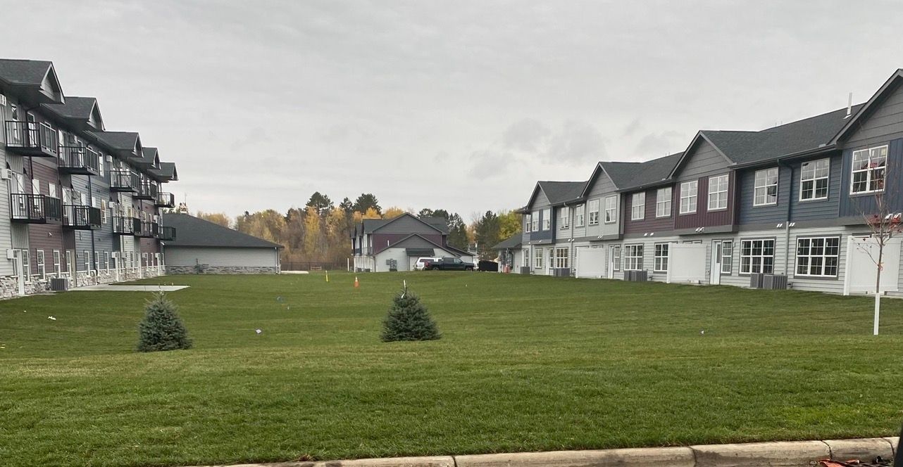 A green lawn sits between two multi-story apartment buildings with gray siding and gabled roofs under an overcast sky.