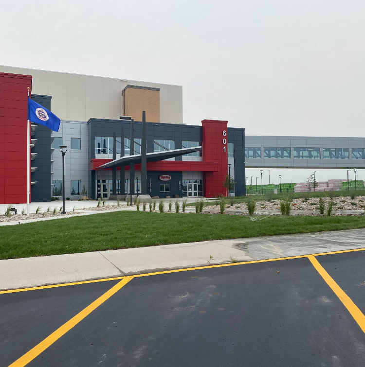 A modern office building with red and dark gray siding, a flag, and a glass-enclosed pedestrian bridge overhead.