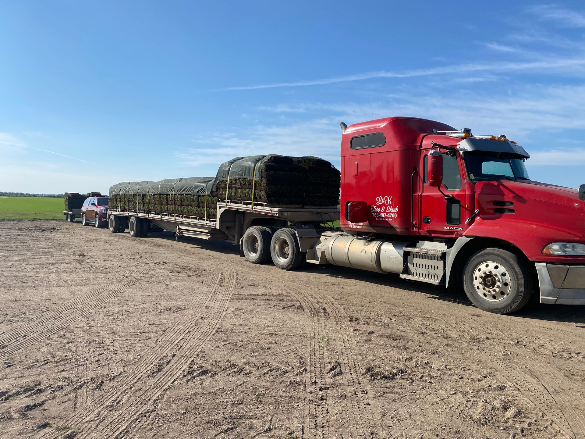 A red semi-truck parked in a field, hauling a flatbed trailer loaded with large round hay bales under a clear blue sky.