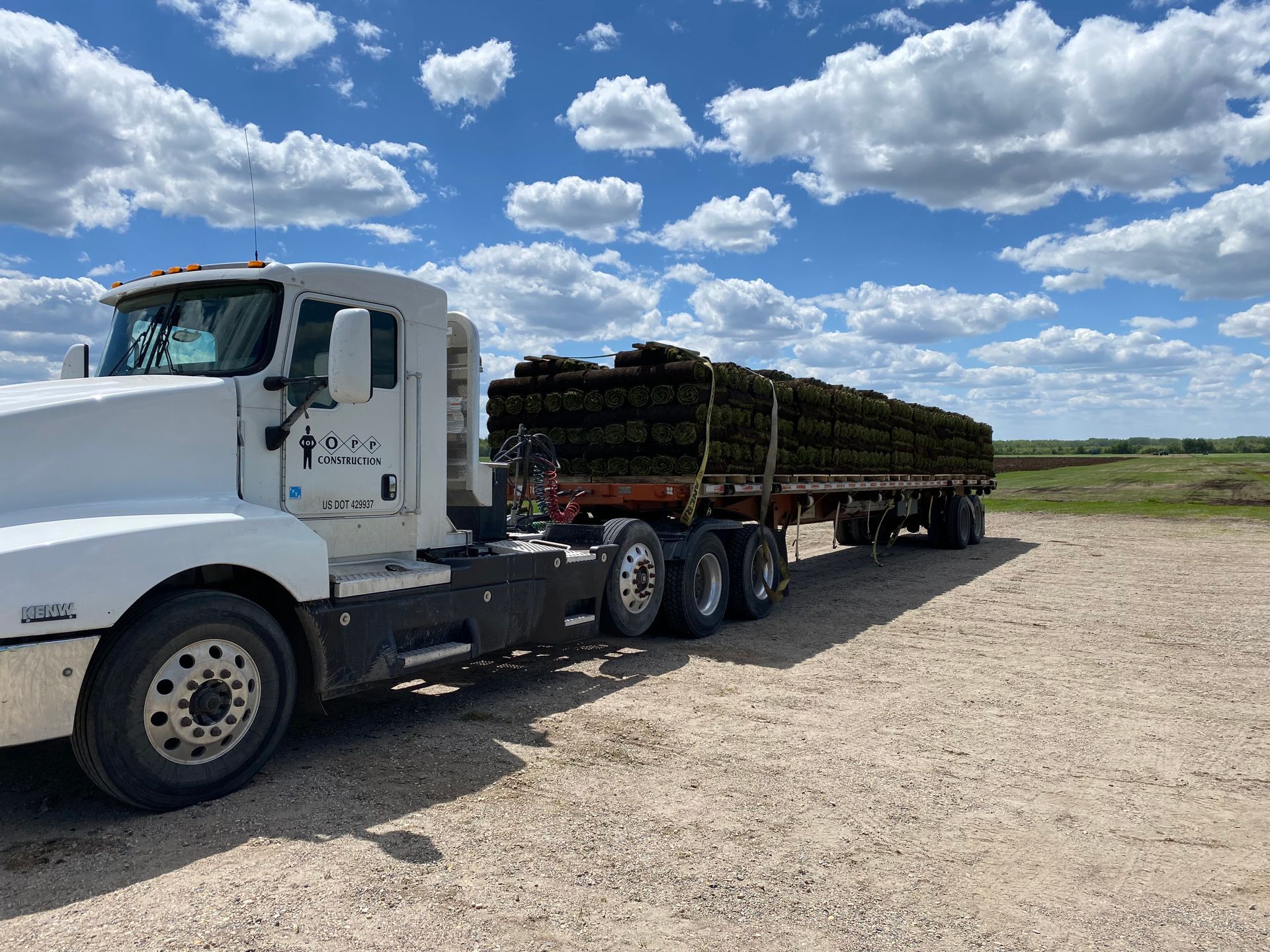 A white semi-truck hauls a flatbed trailer stacked with rolls of sod on a gravel lot under a bright, cloudy blue sky.