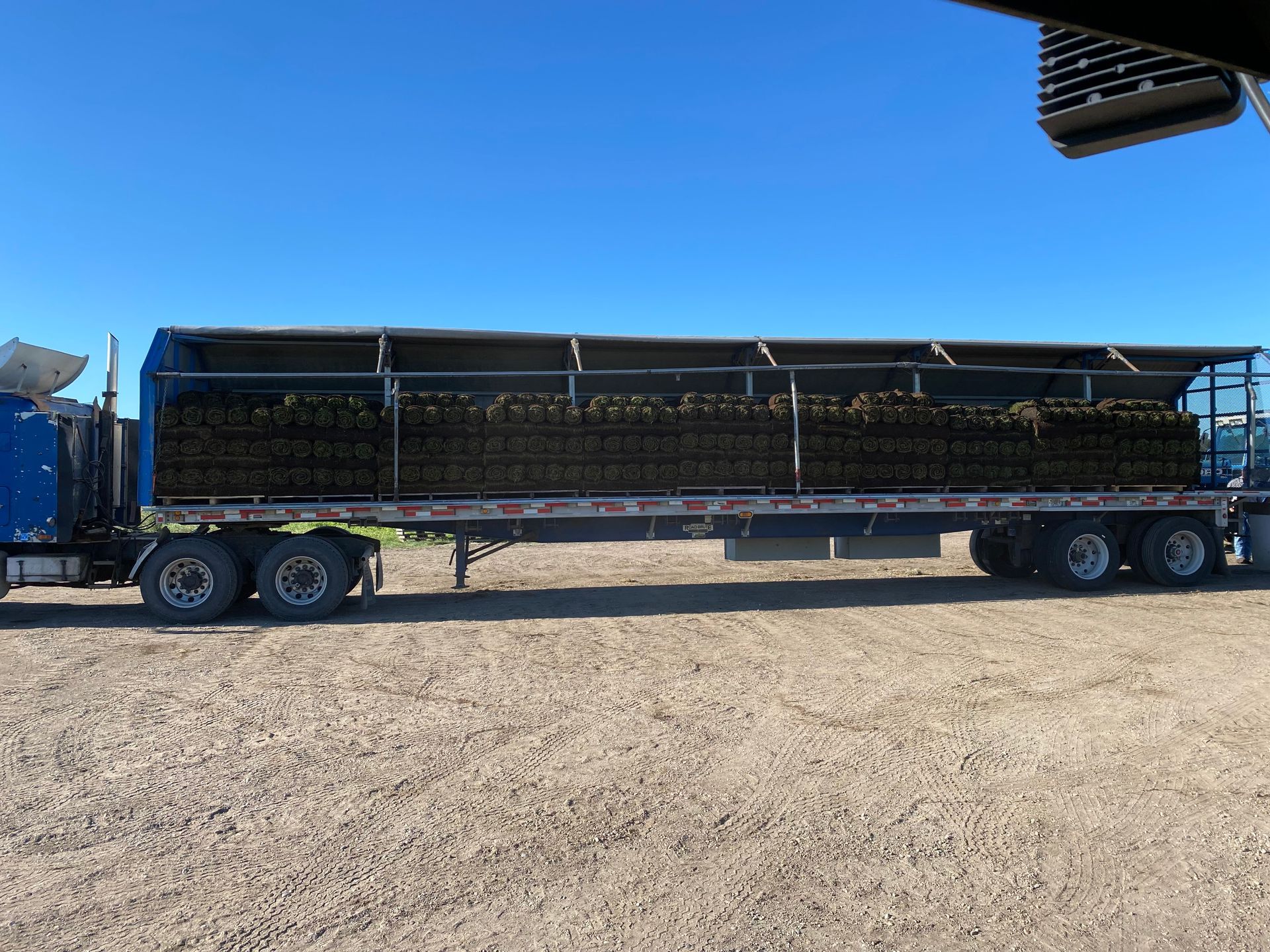 A side view of a semi-truck trailer filled with stacked dark cylindrical hay bales against a bright blue sky.