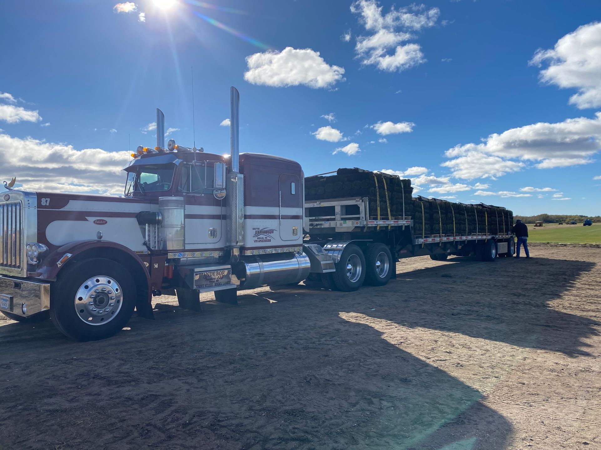 A maroon and white semi-truck with a flatbed trailer loaded with dark cargo parked on a dirt lot under a sunny blue sky.