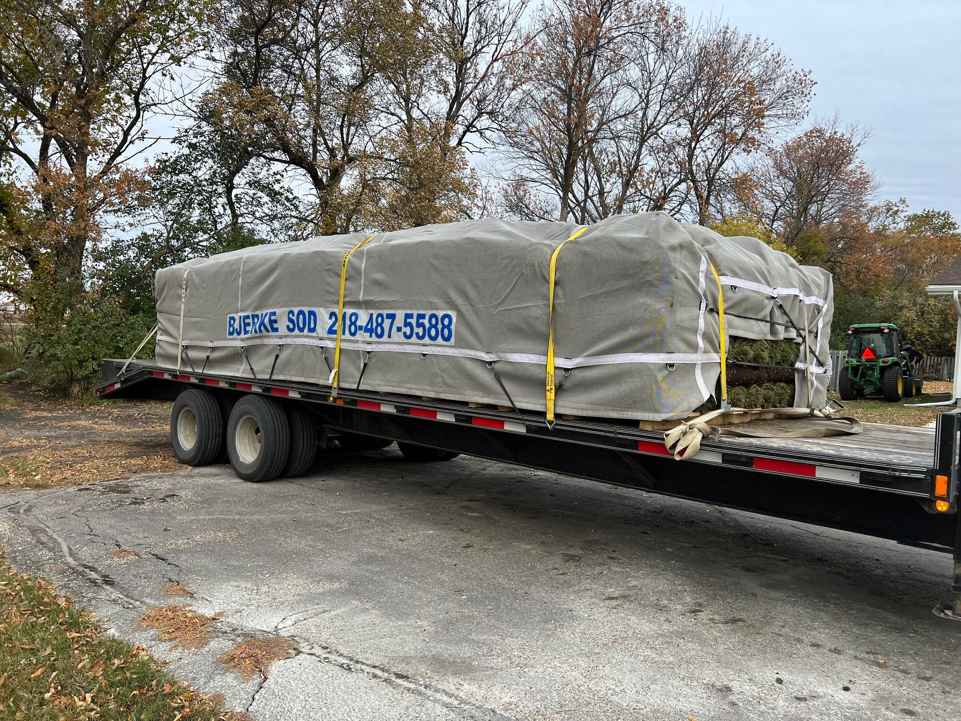 A flatbed trailer carrying a large, grey-covered load with a phone number printed on the side, parked on an asphalt lot.