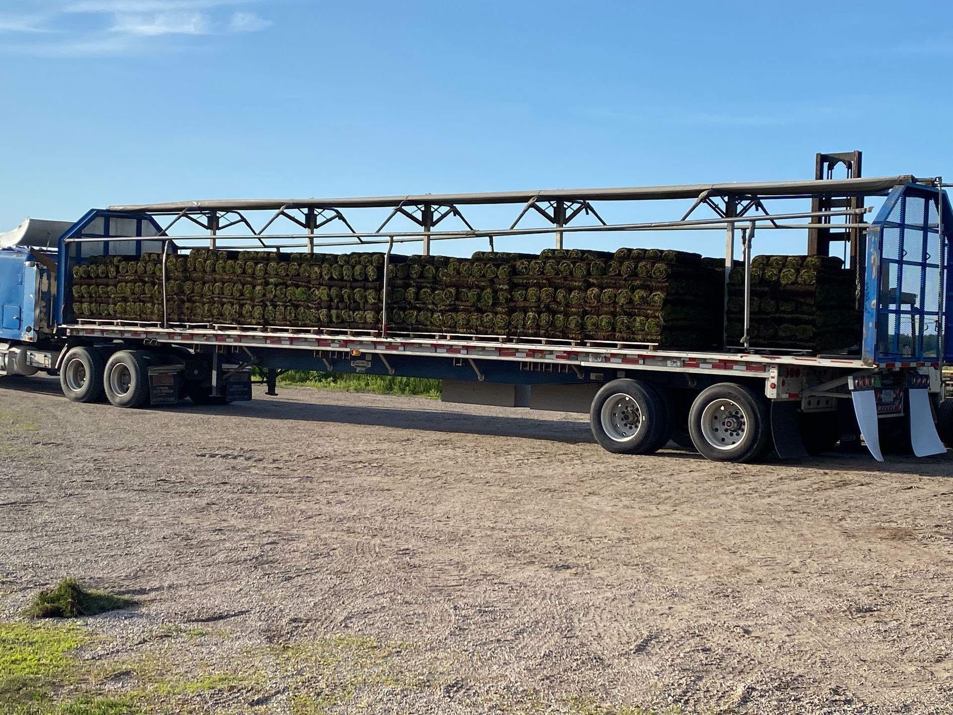 A flatbed semi-truck loaded with rectangular pallets of turf, parked on a gravel surface under a clear blue sky.