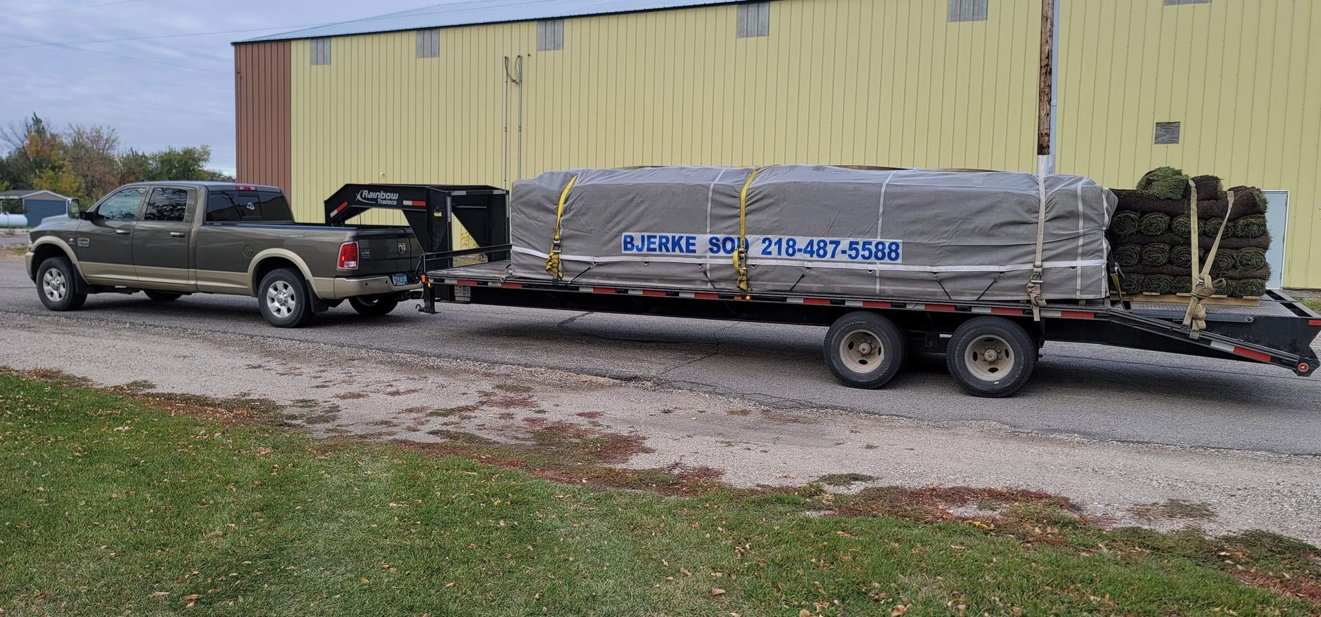 A two-tone Ford pickup truck pulling a gooseneck trailer loaded with a large, tarp-covered pallet in front of a yellow shed.