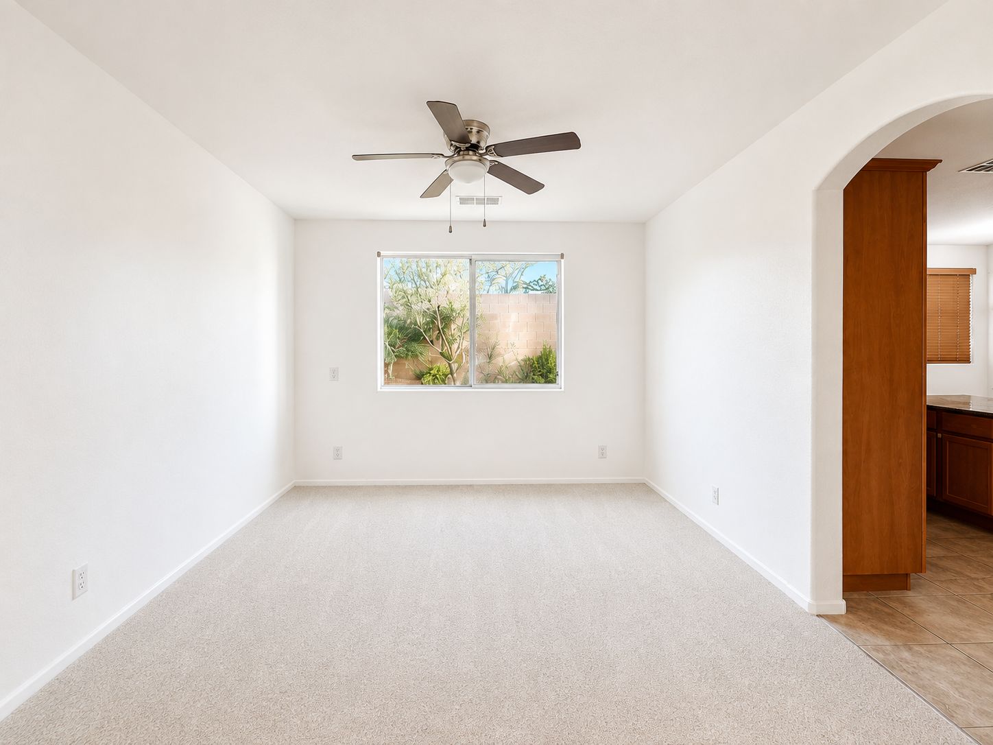 Empty white living room with ceiling fan, window, carpet, and doorway to kitchen