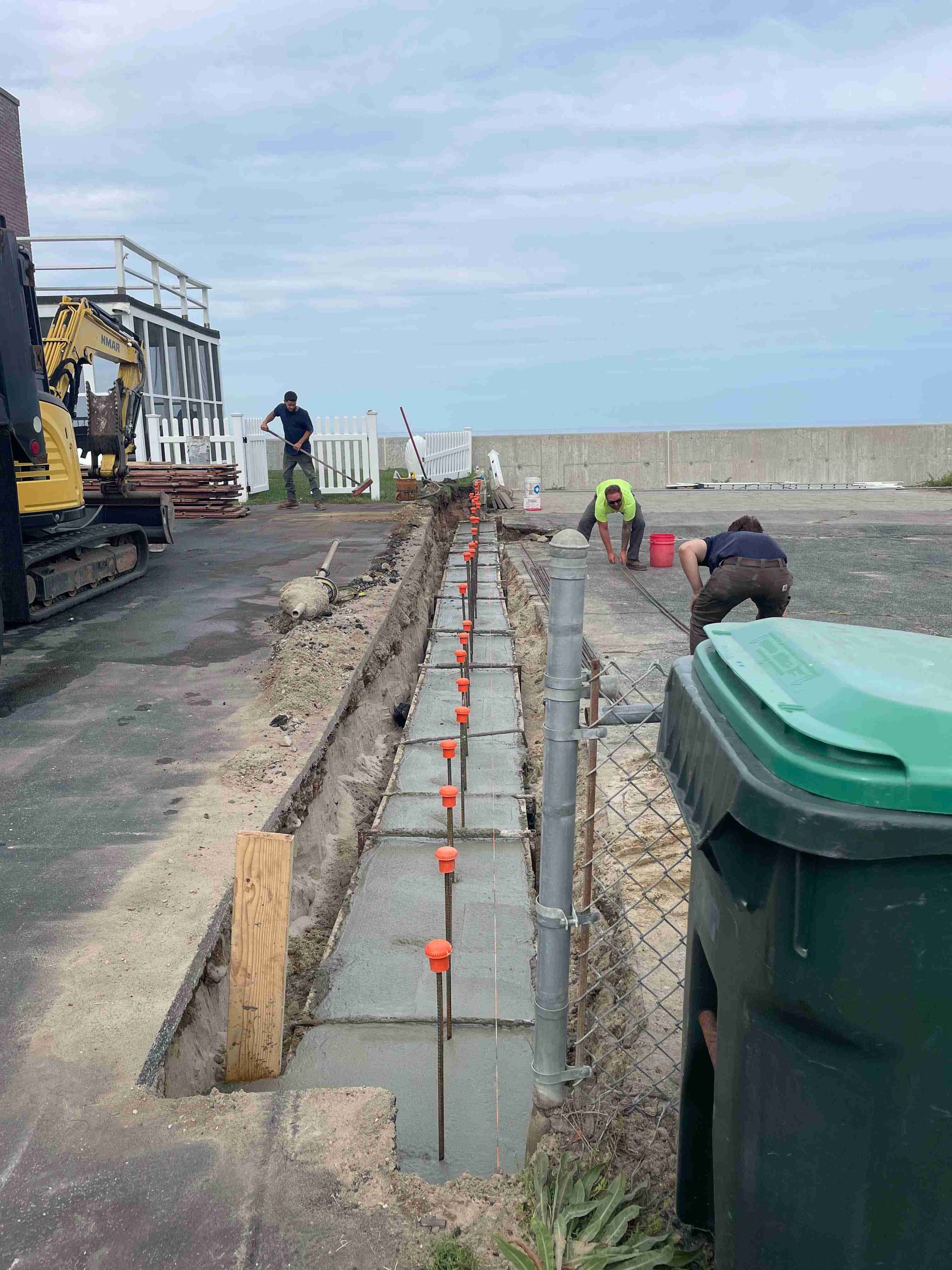 Coastal construction site with a long concrete trench, workers, and equipment beside a road and sea.