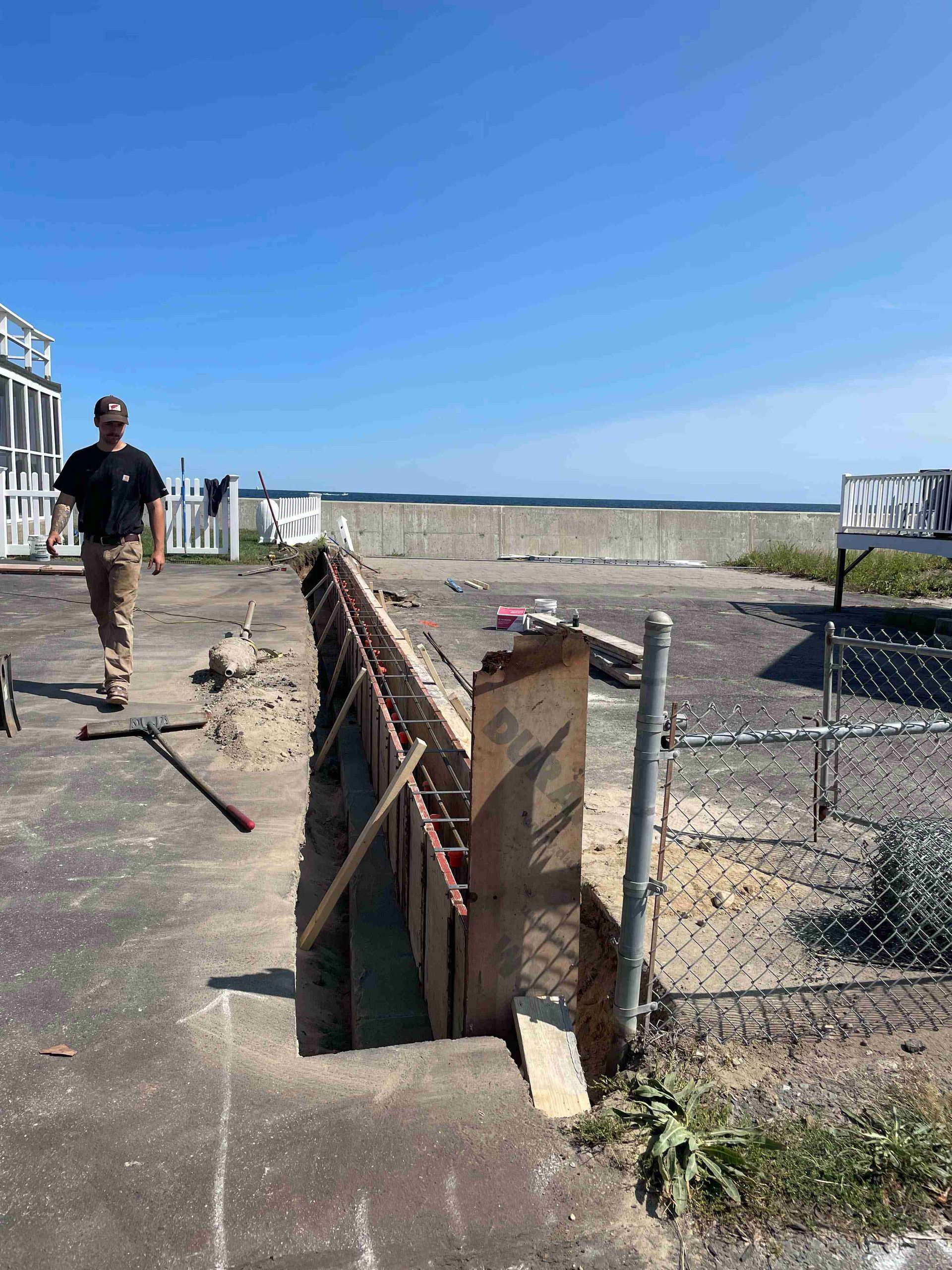 Man walking on a construction site beside a brick wall and chain-link fence under a clear blue sky