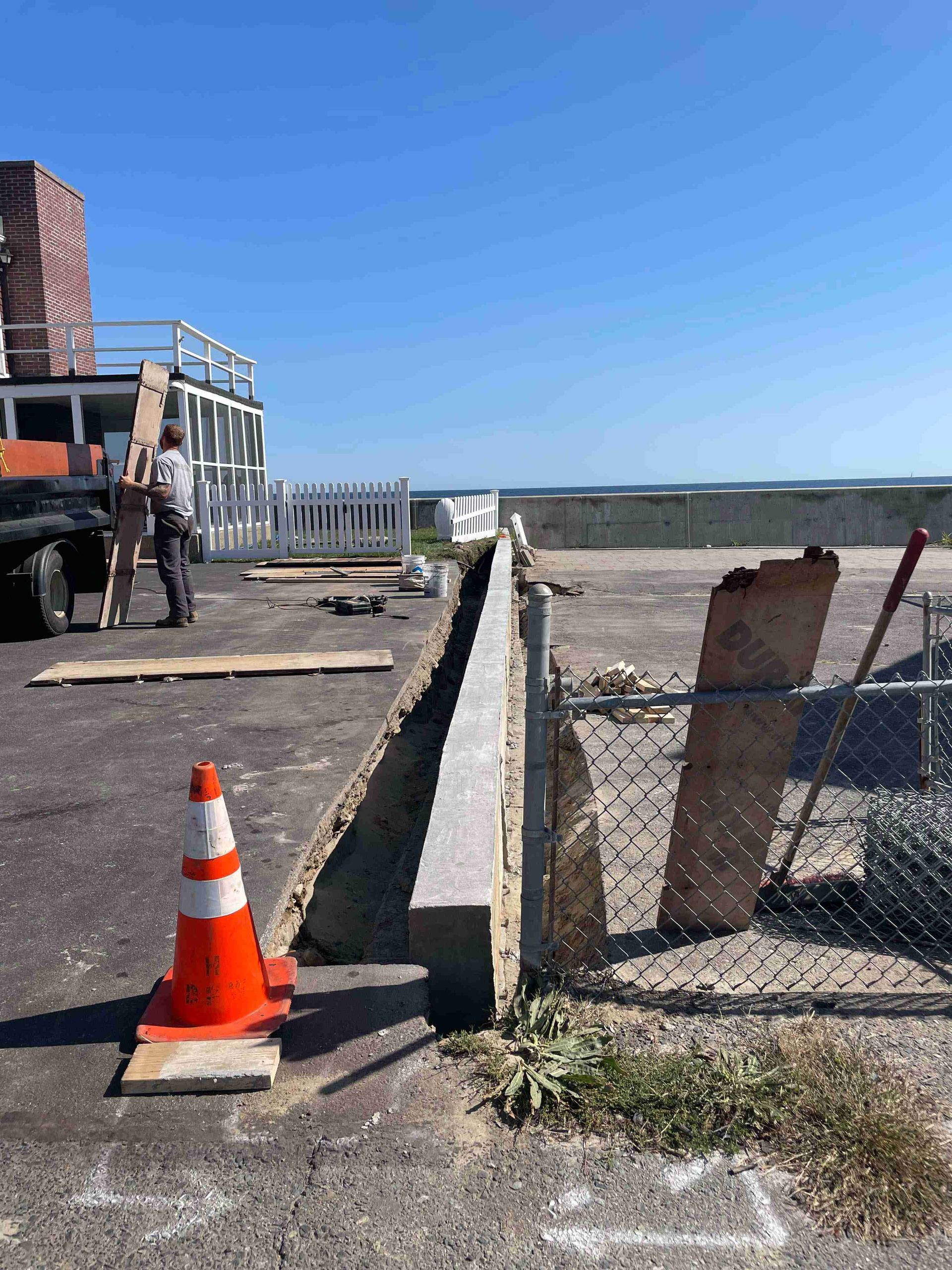 Construction site beside a building with an orange cone, fence, and worker near equipment on a sunny day