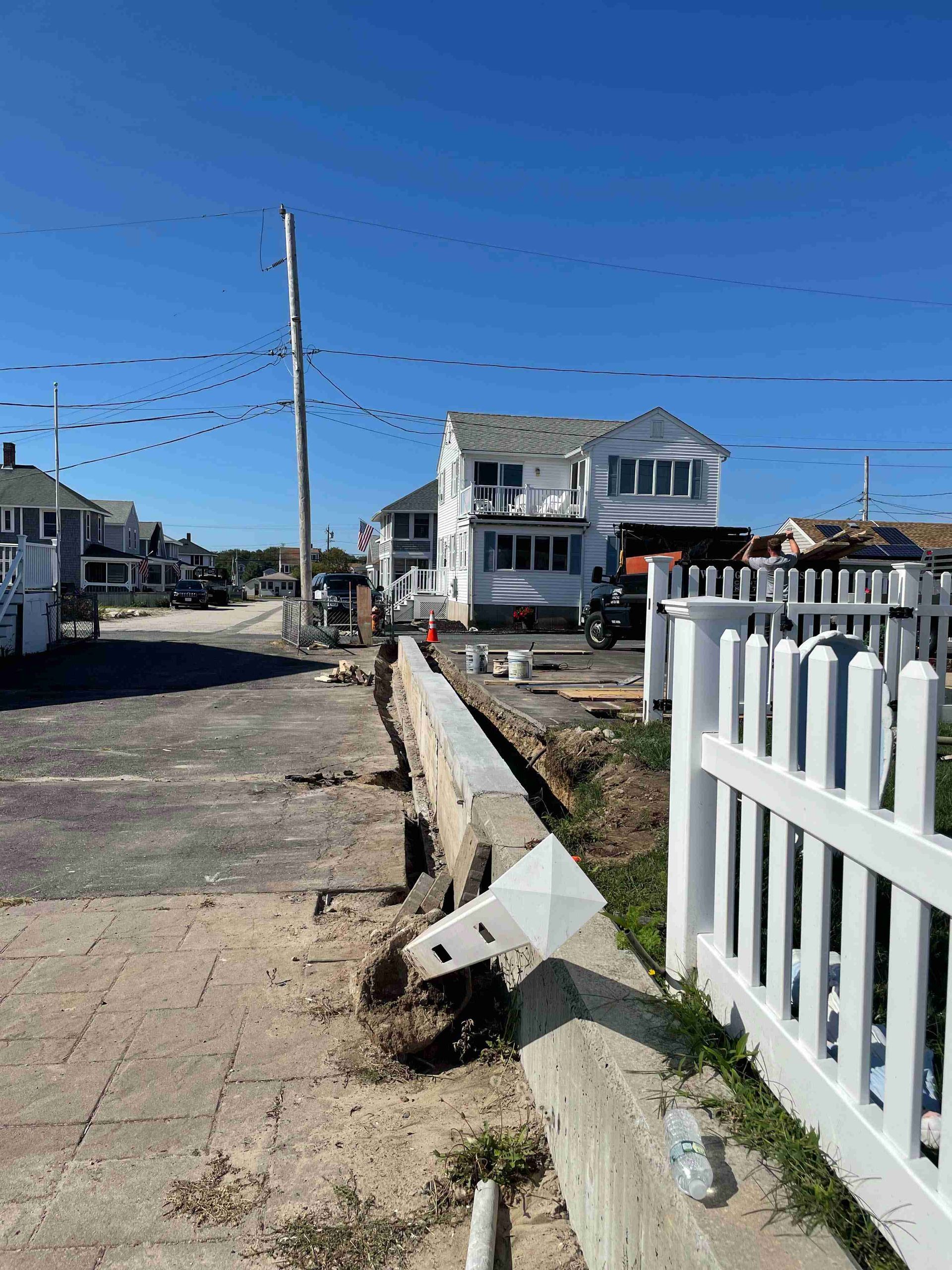 Quiet suburban street with a white two-story house, fence, utility pole, and clear blue sky