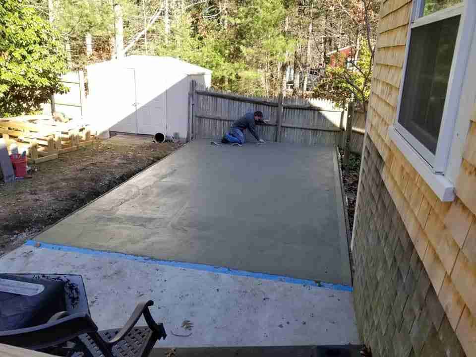 Concrete patio beside house with a blue tarp-covered pile near the fence and trees beyond